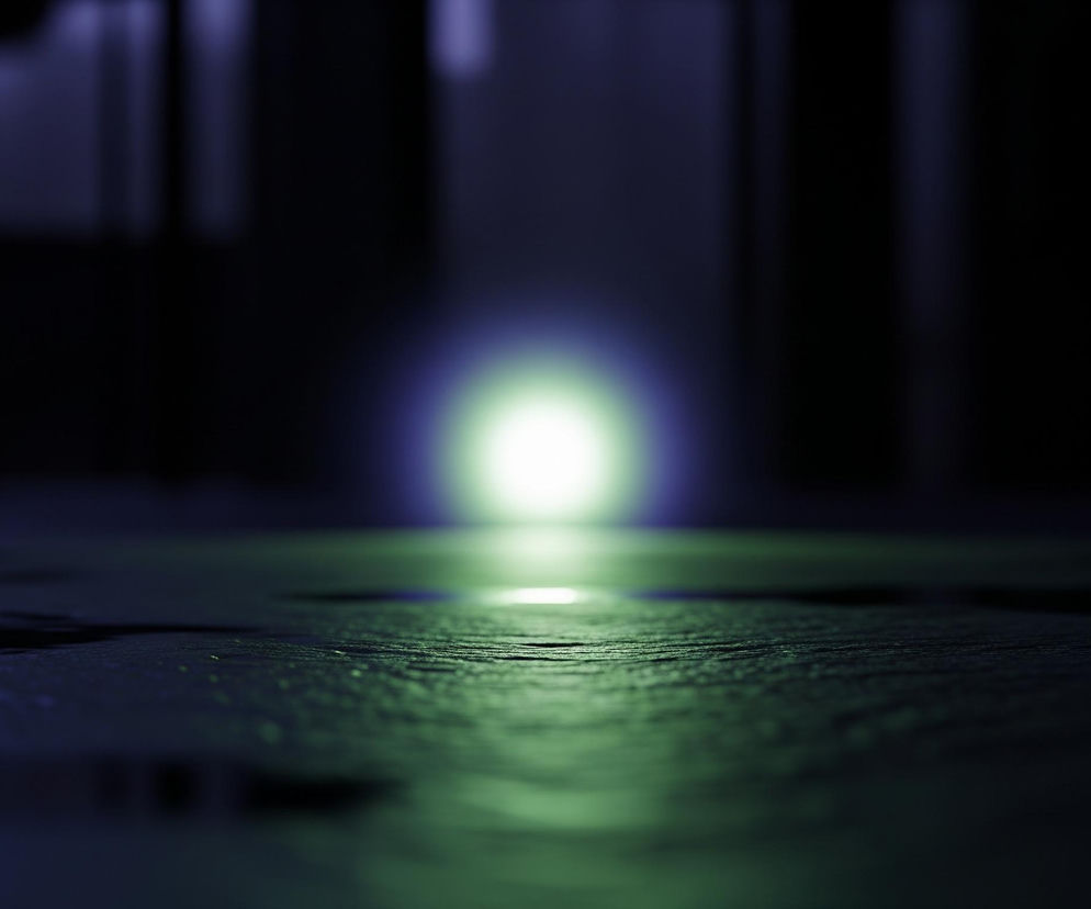 Na ciemnym, rozmytym tle widać intensywne, jasne światło w odcieniach zieleni i fioletu, które stanowi centralny punkt kompozycji. Światło to odbija się na nierównej, mokrej powierzchni przypominającej asfalt, zajmującej dolną połowę kadru. Powierzchnia jest porysowana i pokryta kałużami, z zielonkawym odcieniem w miejscu odbicia światła, przechodzącym w ciemny granat w dalszych planach. Tło jest bardzo ciemne, z nieostrymi, pionowymi smugami w odcieniach ciemnego fioletu, sugerującymi sylwetki budynków lub innych struktur.