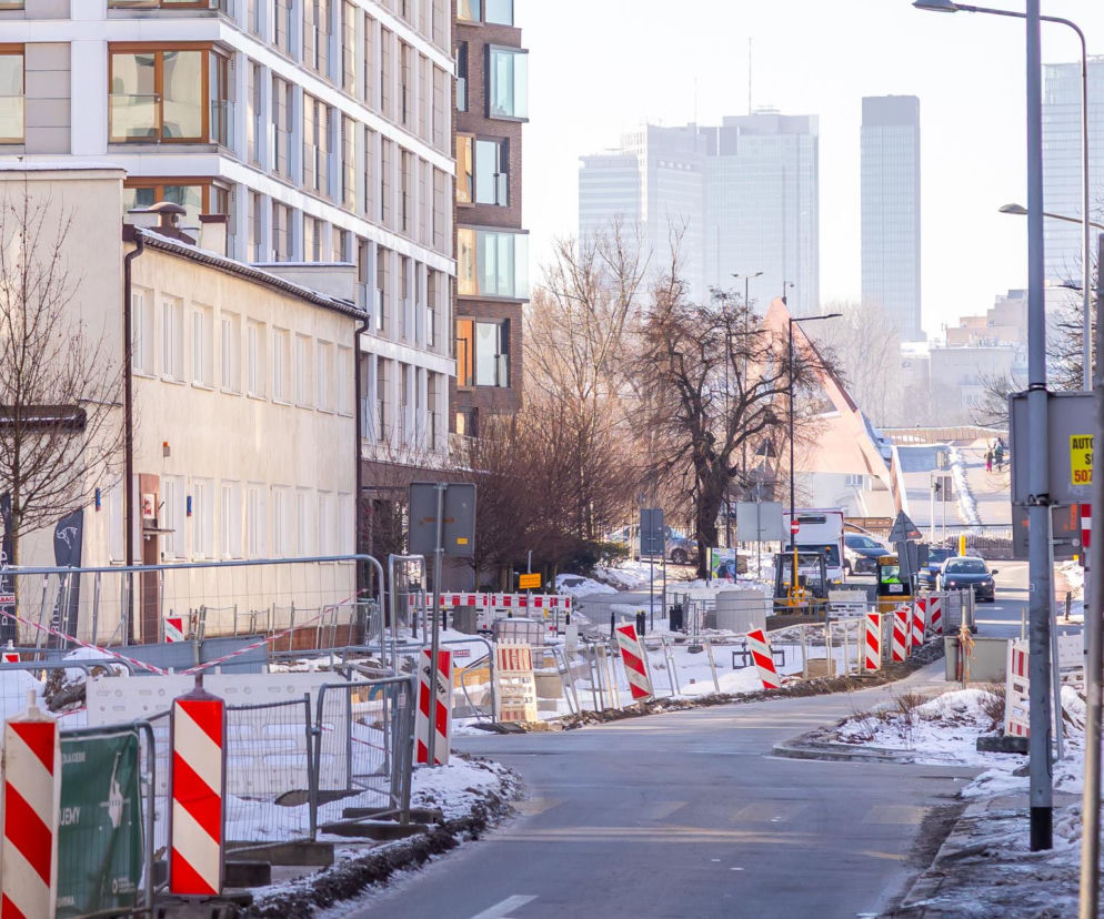 Ulica Okrzei w Warszawie zimą, z widocznymi elementami placu budowy: ogrodzeniami i znakami drogowymi. Po prawej piesza z torbą, a w tle nowoczesne budynki. Prace na ul. Okrzei, o których przeczytasz na Architektura Murator Plus, mają przekształcić ją w przestrzeń przyjazną dla pieszych i rowerzystów.