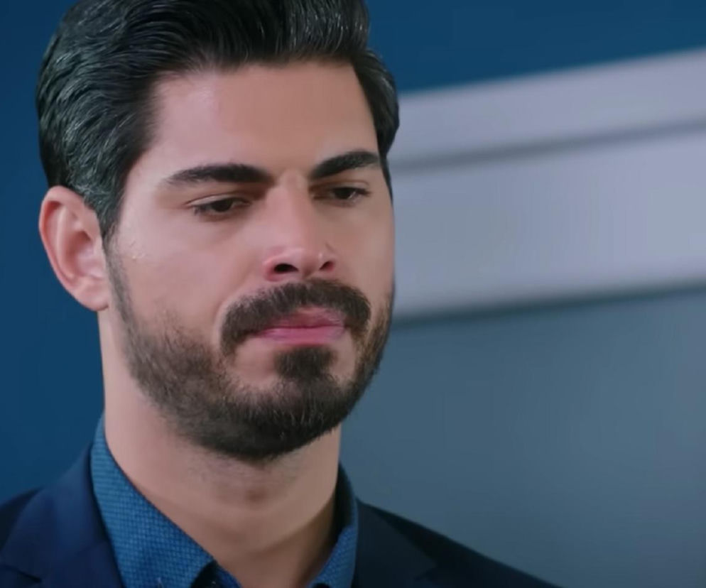 Mężczyzna z brodą i wąsem, ubrany w granatową marynarkę i niebieską koszulę, spogląda w dół z zamyśloną miną, w tle błękitna ściana. Zdjęcie przedstawia Cihana z serialu Panna młoda, o którym przeczytasz więcej na portalu Super Seriale.