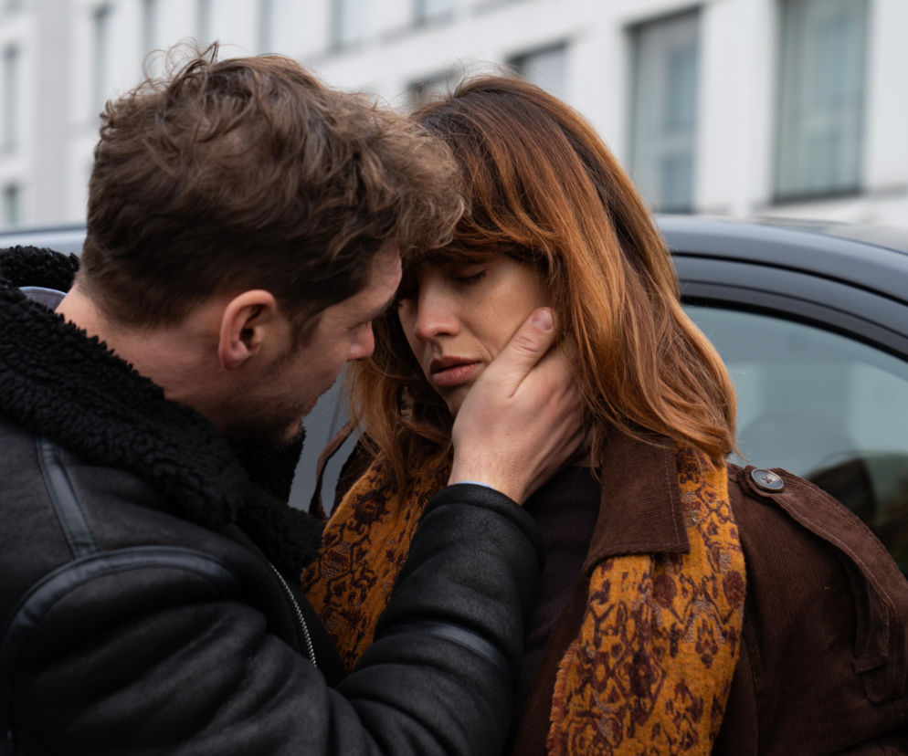 Zbliżenie na Marcina Chodakowskiego (Mikołaj Roznerski) i Kamę (Michalina Sosna) z serialu „M jak miłość”. Marcin delikatnie obejmuje twarz Kamy, która ma zamknięte oczy i smutny wyraz twarzy. Oboje stoją blisko siebie, opierając czoła. W tle widoczny jest fragment czarnego samochodu i jasnego budynku. O tym, jak Marcin ulegnie Kamie, by ratować małżeństwo, możesz przeczytać na portalu Super Seriale.