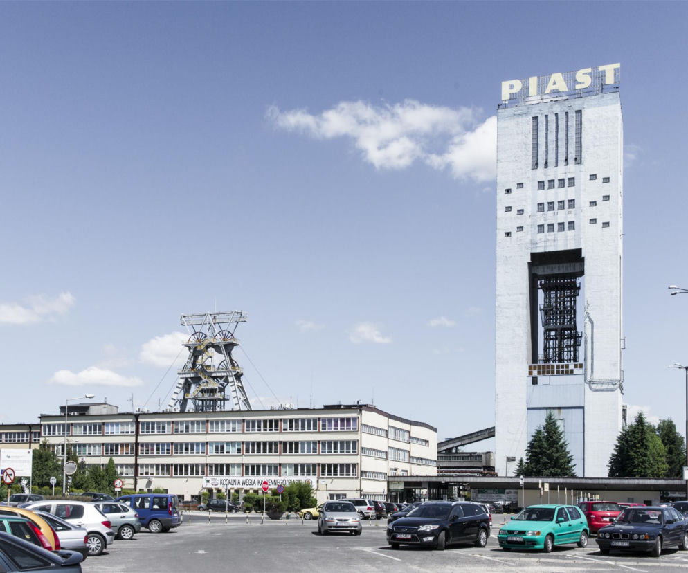 Kopalnia węgla kamiennego Piast w Bieruniu, z charakterystycznym szybem i budynkami biurowymi, otoczona parkingiem z samochodami pod błękitnym niebem. W tle widoczna wieża wyciągowa i zieleń drzew. Planowane zagospodarowanie terenów pokopalnianych to ważny temat na Super Biznes.