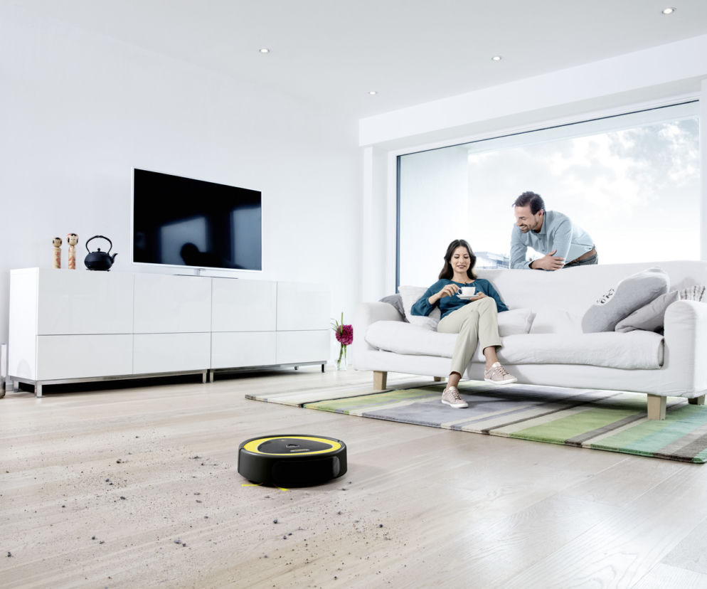 Robot sprzątający odkurza jasną podłogę w salonie. W tle para odpoczywająca na sofie. O aranżacji wnętrz czytaj na Urządzamy.