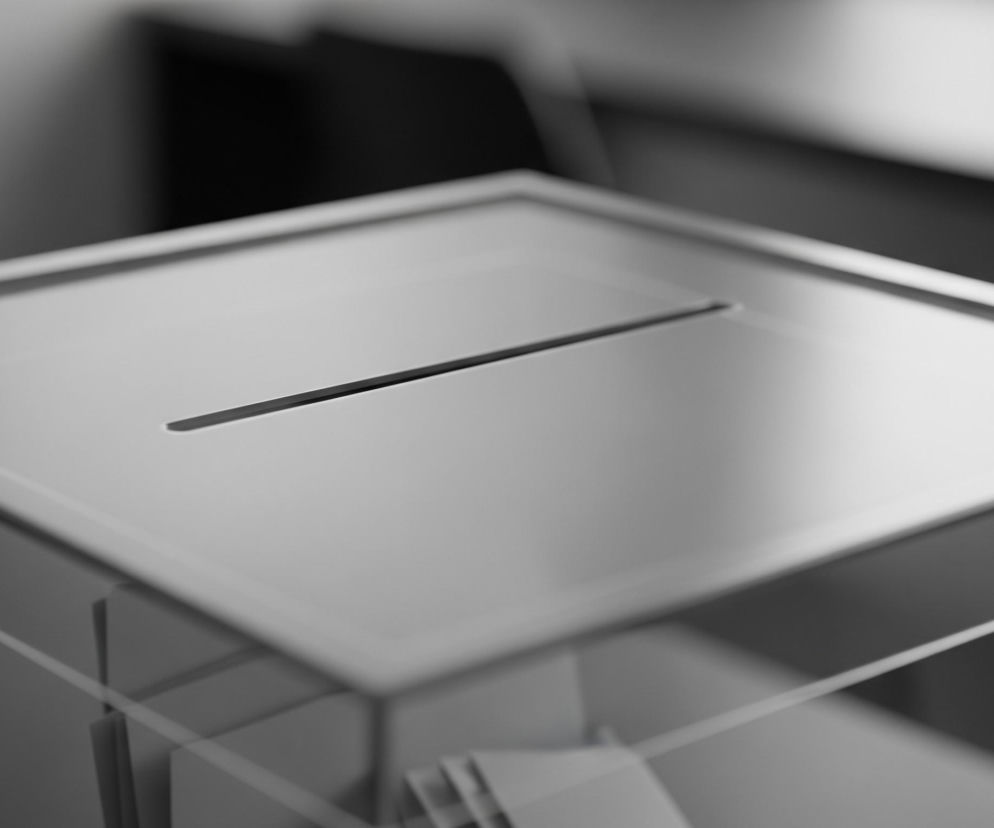 Przejrzysta urna wyborcza z pleksiglasu, lekko przechylona, dominują odcienie szarości. W górnej części widoczny jest wąski, poziomy otwór na karty do głosowania. Wewnątrz urny widać kilka złożonych białych papierów, prawdopodobnie kart wyborczych. Tło jest rozmyte, z niewyraźnymi ciemnymi i jasnymi obszarami.