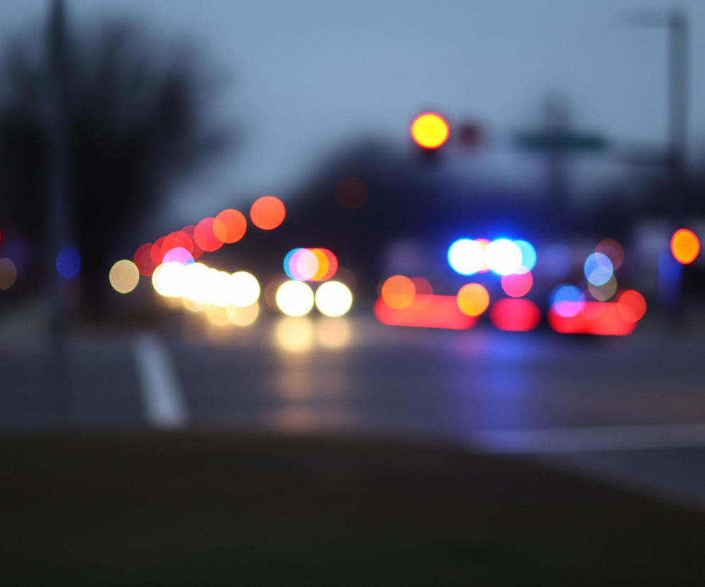 Rozmyty obraz przedstawia nocną scenę uliczną z dominującymi światłami samochodów i sygnalizacji świetlnej. W centralnej części widoczne są intensywne, niebiesko-czerwone światła sygnalizacyjne, które sugerują obecność pojazdu uprzywilejowanego. Po lewej stronie drogi rozmieszczone są rzędy żółtych i pomarańczowych świateł, a nieco w oddali widoczne jest pojedyncze, jasne żółte światło. Ciemny, szarobłękitny nieboskłon stanowi tło dla rozmytych kształtów drzew i słupów, a u dołu obrazu znajduje się ciemna, niewyraźna nawierzchnia drogi z pojedynczym, białym pasem.