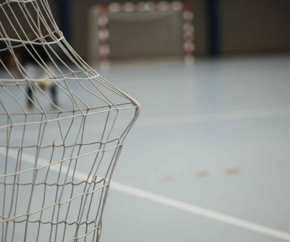 Prawa część siatki bramki sportowej, wykonana z jasnych sznurków tworzących kwadratowe oczka, dominuje lewą stronę kadru. Za nią widać jasno-niebieskie boisko z białą linią i kilkoma rozmytymi, pomarańczowymi punktami. W tle, po prawej stronie boiska, widoczne są rozmyte sylwetki dwóch osób w ciemnych strojach, jedna z nich ma zielone obuwie. Na dalszym planie, pośrodku, znajduje się niewyraźna bramka z czerwono-białymi pasami.