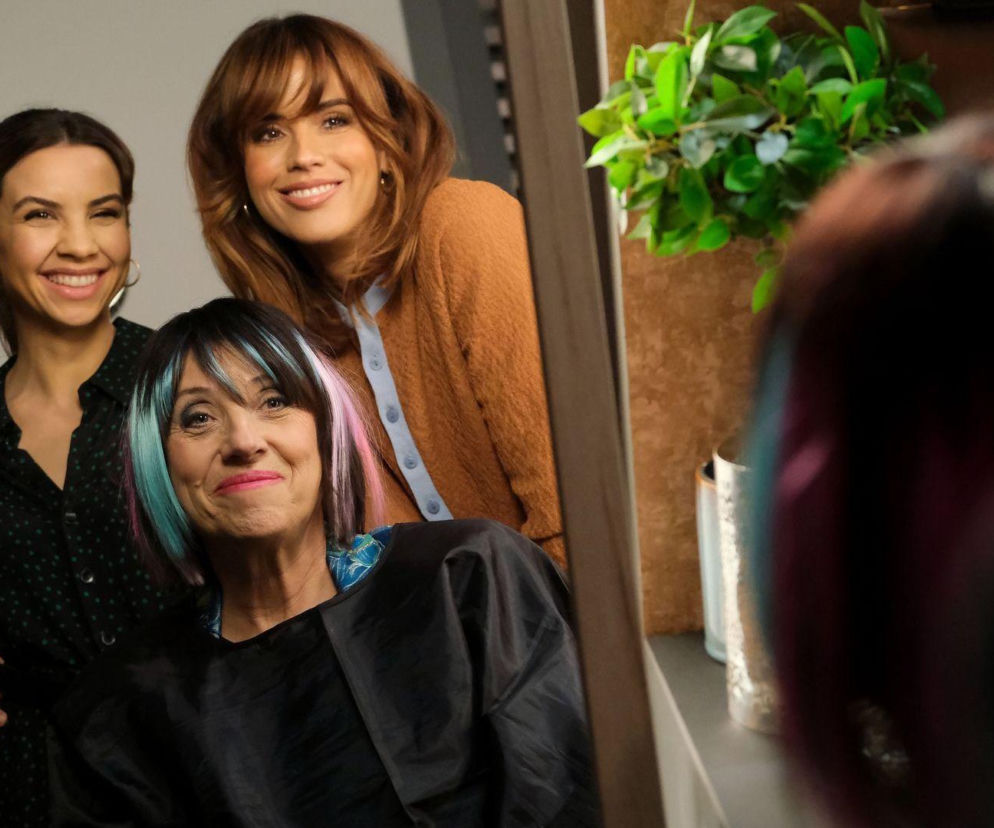 Iwona Kryńska z M jak miłość z awangardową, kolorową fryzurą, siedzi przed lustrem w salonie fryzjerskim. Za nią uśmiechają się Kama i Aneta, zmartwione jej metamorfozą. Scena odzwierciedla dramatyczne wydarzenia z serialu dostępne na Super Seriale.