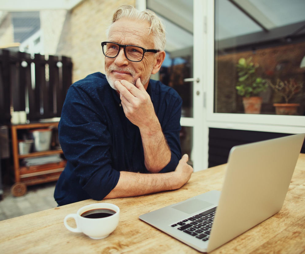 Starszy mężczyzna z siwą brodą i w okularach, z filiżanką kawy i laptopem na drewnianym stole, zamyślony patrzy w bok. Zdjęcie ilustruje temat wpływu kawy na zdrowie serca i arytmię, o czym przeczytasz więcej na Poradnik Zdrowie.