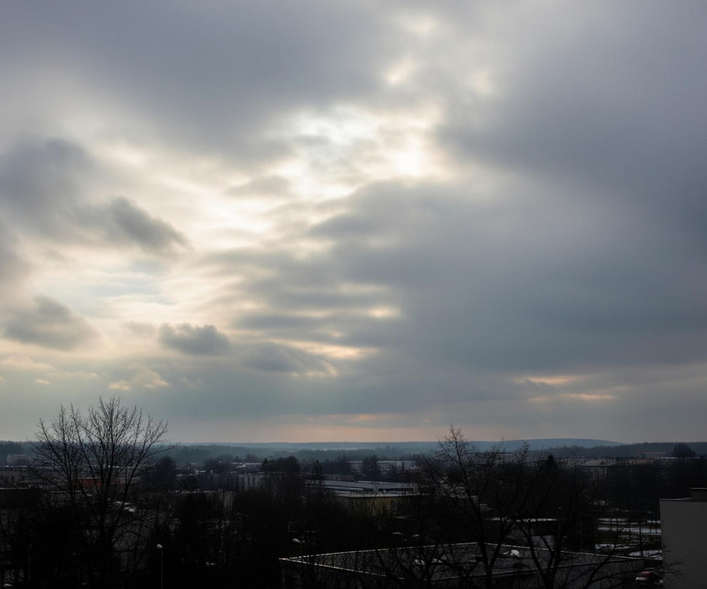 Pochmurne niebo z prześwitami słońca, wypełniające dwie trzecie górnej części kadru, dominuje nad widokiem miejskim. Chmury mają odcienie szarości, od ciemnego grafitu po jaśniejsze, niemal białe fragmenty w miejscach, gdzie przebija się światło. Dolna jedna trzecia obrazu przedstawia panoramę miasta w odcieniach szarości i czerni, z ciemnymi sylwetkami budynków i bezlistnych drzew. Horyzont tworzy linię lasu lub wzniesień, a w oddali widać subtelne, różowo-pomarańczowe zabarwienie nieba tuż nad nim.