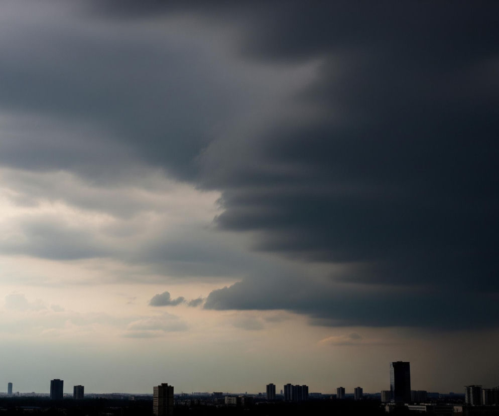 Panoramę miejską pod dramatycznym, burzowym niebem. Górna połowa zdjęcia zdominowana jest przez ciemne, ciężkie chmury, które przechodzą od jasnoszarego na lewej stronie do niemal czarnego na prawej, tworząc wyraźne, dynamiczne przejście. Poniżej chmur, na horyzoncie, widoczne są sylwetki licznych budynków, głównie wieżowców i mniejszych bloków, które w większości są zacienione i wyglądają na czarne kształty na tle jaśniejszego nieba. Niebo w dolnej części obrazu, tuż nad zabudowaniami, ma jaśniejszy, mleczno-szary odcień, co sugeruje zbliżający się lub oddalający opad.