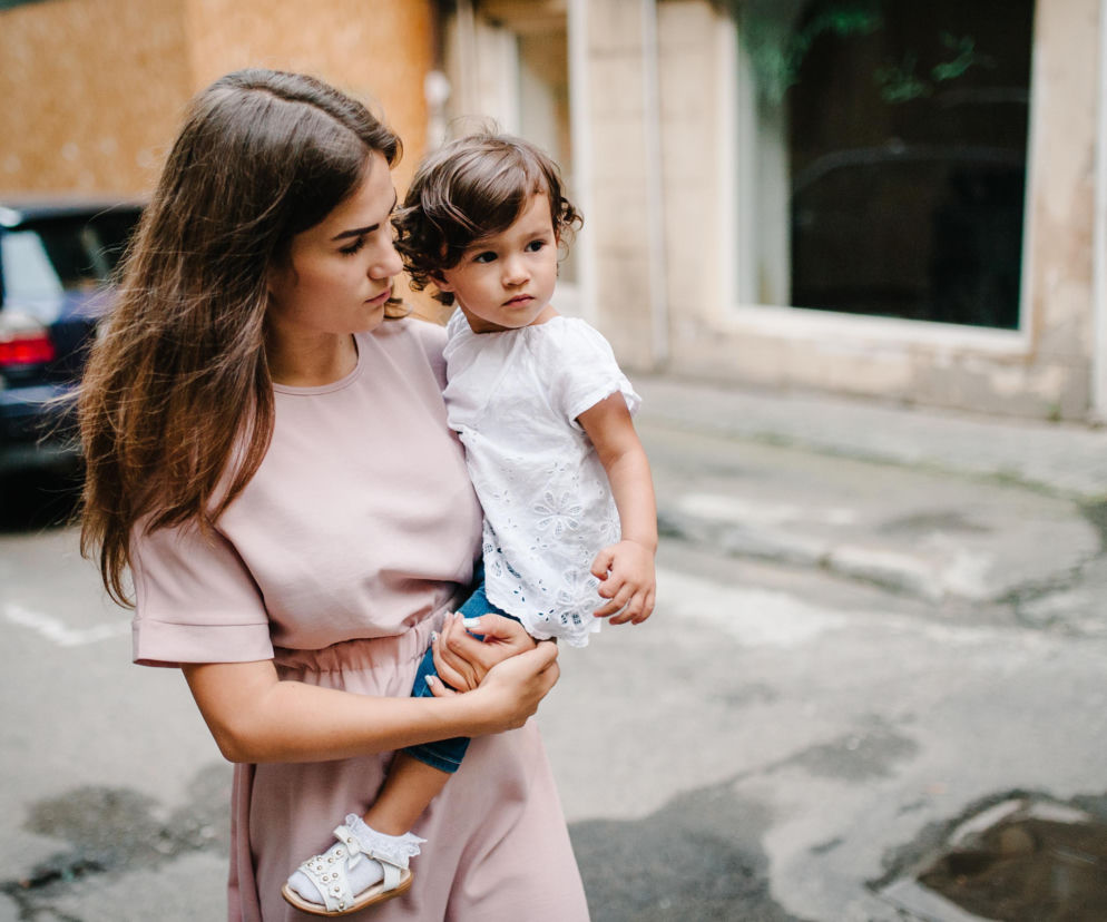 Kobieta w jasnoróżowej sukience, niosąca dziecko na biodrze, spogląda w prawą stronę. Dziecko w białej bluzce z haftem i dżinsach, z bujnymi kręconymi włosami, również patrzy w bok. Tło to ulica z budynkami, a w oddali widać samochód, co ilustruje artykuł o prawidłowym noszeniu dziecka, by nie obciążać kręgosłupa, dostępny na portalu Poradnik Zdrowie.