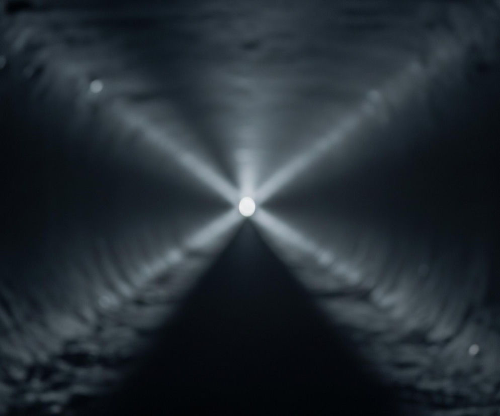 Ciemny, cylindryczny tunel z rozmytą teksturą na ścianach i suficie jest oświetlony jasnym, białym punktem światła w oddali, znajdującym się dokładnie w centrum obrazu. Światło tworzy rozchodzące się promienie, które przecinają się w kształcie litery X, rozjaśniając otoczenie i nadając mu głębię perspektywy. Otoczenie tonie w odcieniach szarości i czerni, a nieliczne rozproszone jasne punkty sugerują odbicia lub dodatkowe źródła światła, które są poza ostrością.