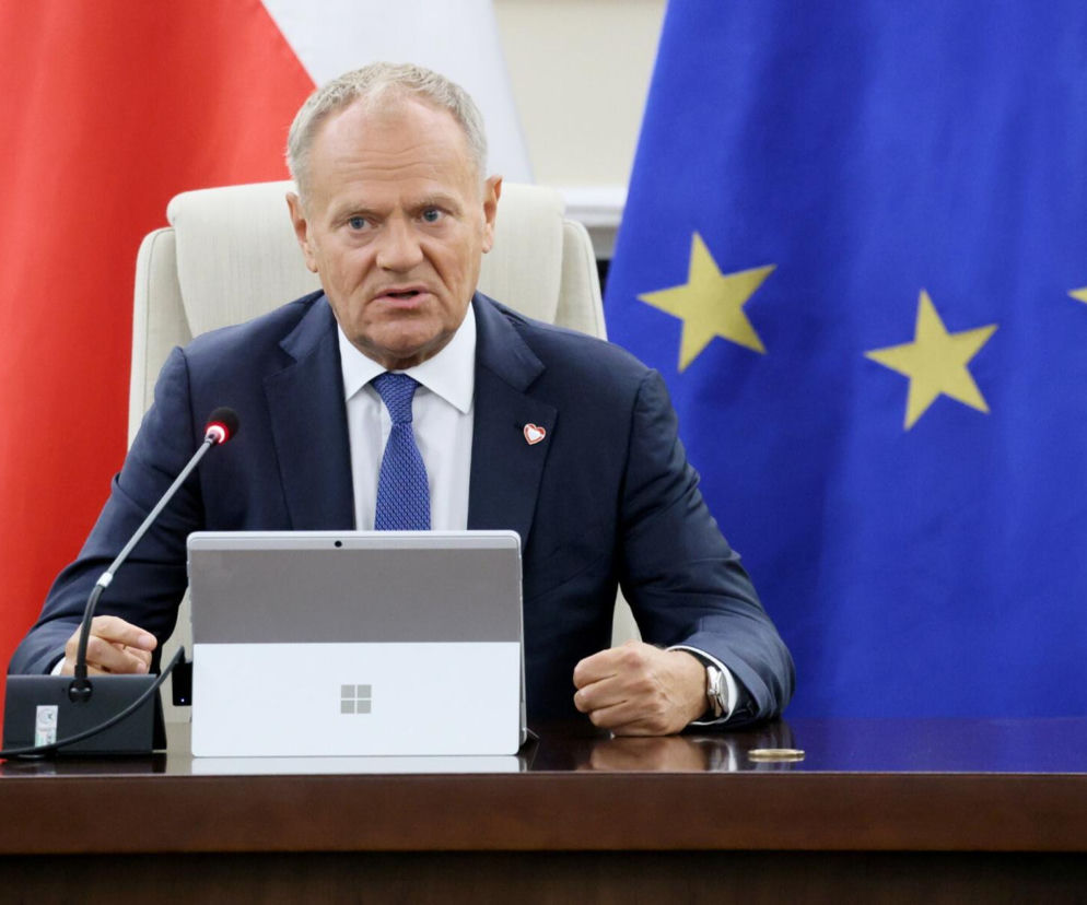Premier Donald Tusk przy stole z laptopem na tle flag Polski i Unii Europejskiej. O nowym podatku czytaj na SE Superbiz.