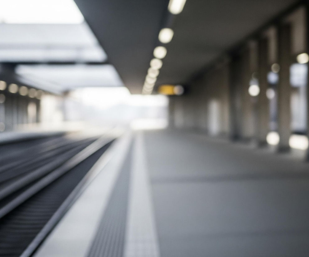 Długi peron kolejowy rozciąga się w prawo, a tory kolejowe wiodą w głąb obrazu po lewej stronie. Na pierwszym planie widoczne są szare tory, rozmazane, przechodzące w niewyraźny horyzont. Sufit peronu jest podparty rzędem prostokątnych słupów po prawej stronie, a białe, podłużne lampy rozmieszczone są w regularnych odstępach zarówno nad peronem, jak i nad torami, tworząc rozmyte punkty światła. Całe ujęcie jest nieostre, dominują w nim odcienie szarości i bieli, z pojedynczym żółtym elementem widocznym w oddali, pod sufitem.