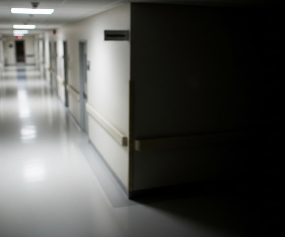 Wąski, jasny korytarz rozciąga się w głąb obrazu, zlewając się z oddaleniem. Białe ściany i jasna podłoga odbijają światło z podłużnych lamp sufitowych. Po obu stronach korytarza widoczne są drzwi do pomieszczeń, a nad nimi wiszą ciemne tabliczki informacyjne z niewyraźnymi napisami. Po prawej stronie, na pierwszym planie, ściana gwałtownie przechodzi w ciemność, tworząc mocny kontrast i sugerując kąt lub zacieniony obszar.