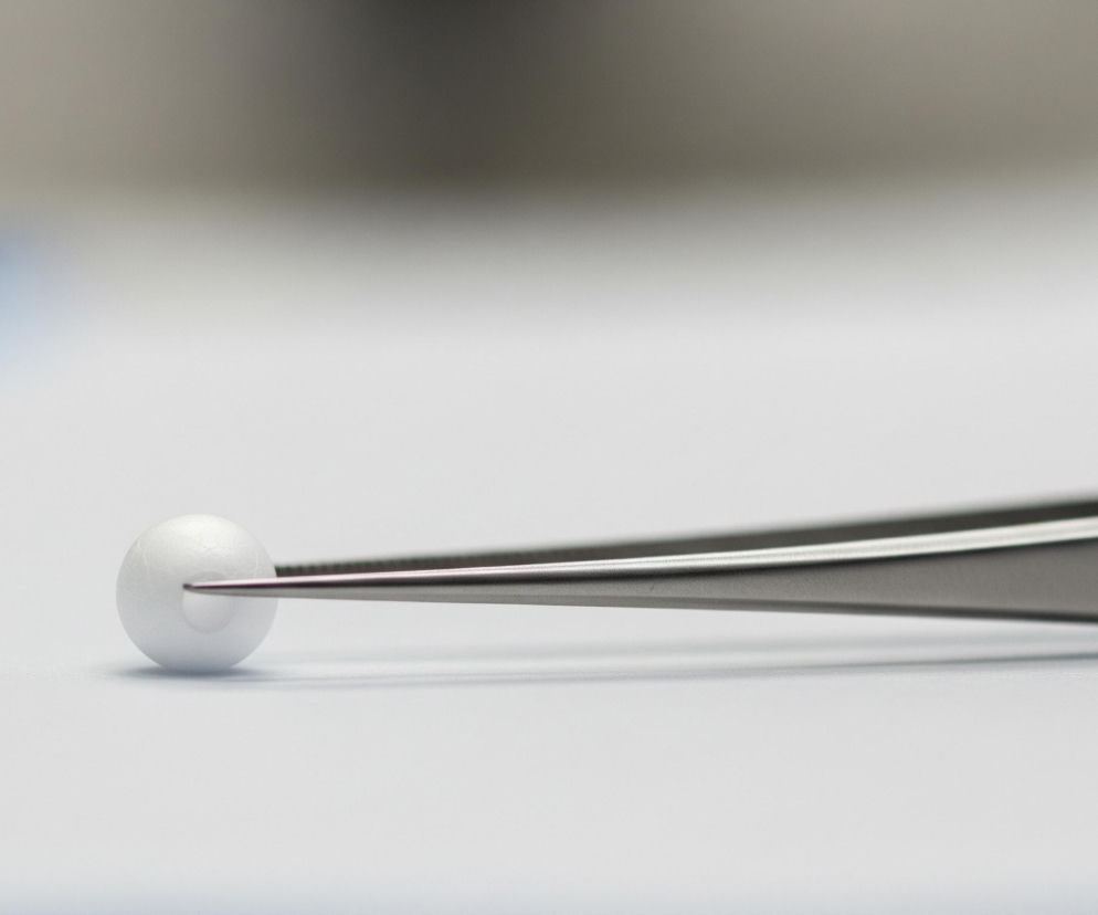 Centralnie umieszczona biała, kulista kuleczka jest chwytana za pomocą metalowej pęsety o szpiczastych końcówkach. Pęseta, w odcieniach szarości, rozciąga się od lewej do prawej, z jej prawą stroną lekko rozmazaną, co sugeruje płytką głębię ostrości. Tło jest nieostre, przechodząc od jaśniejszej bieli na dole do ciemniejszych szarości u góry, z subtelnym odcieniem niebieskiego w lewej górnej części.