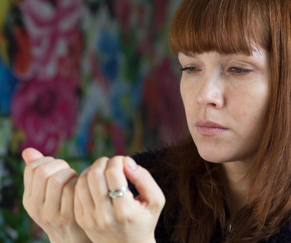 Kobieta z rudymi włosami uważnie oglądająca swoje paznokcie w poszukiwaniu zmian. Na dłoni widoczny pierścionek, w tle rozmazane, kolorowe kwiaty. Sprawdź, co oznaczają linie Beau na paznokciach na Poradnik Zdrowie.