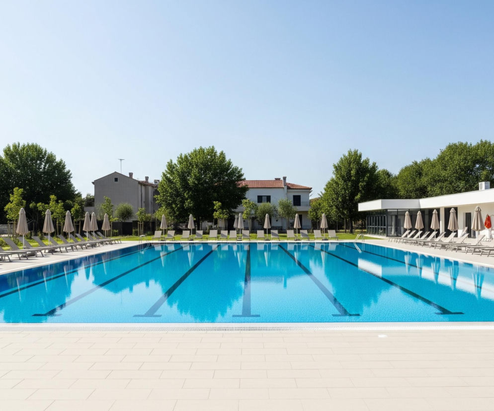 Centralną część obrazu zajmuje duży basen o turkusowej wodzie, podzielony na tory czarnymi liniami na dnie, z jasnym, beżowym brzegiem. Dookoła basenu, na jasnożółtej płytkowej nawierzchni, rozstawione są rzędy szarych leżaków i beżowych parasoli, tworząc strefy wypoczynku po obu dłuższych stronach. W tle widoczne są gęste, zielone drzewa, kilka budynków o jasnych fasadach i czerwonych dachach, a na prawym brzegu basenu znajduje się nowoczesny biały budynek z dużymi oknami, również otoczony leżakami i parasolami.