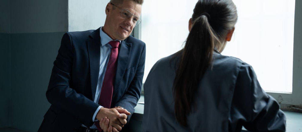 Piotrek (Marcin Mroczek) w eleganckim garniturze i czerwonym krawacie z okularami, uśmiecha się do siedzącej tyłem Magdy (Anna Mucha) z długimi włosami spiętymi w kucyk. Oboje opierają się o parapet w celi więziennej, jasne okno na tle. Więcej o ich losach na Super Seriale.