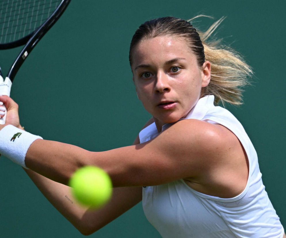 Maja Chwalińska podczas meczu tenisowego, dynamicznie uderzająca piłkę rakietą na korcie trawiastym Wimbledonu. Na ręce widoczna biała opaska z logo Lacoste. O jej występie i relacji z meczu przeczytasz więcej na naszym portalu.