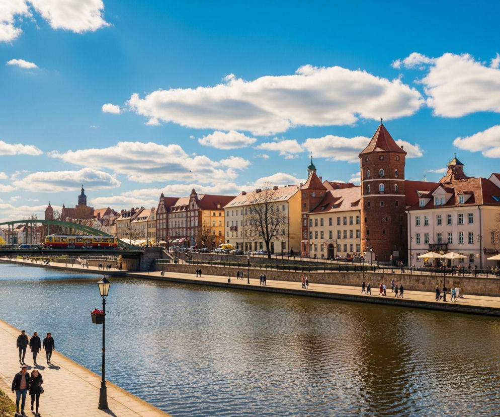 Szeroki kadr ukazuje rzekę z nabrzeżnymi promenadami, wzdłuż których rozciąga się miejska zabudowa pod jasnoniebieskim, bezchmurnym niebem. W oddali, po lewej stronie, widoczny jest zielony most łukowy z przejeżdżającym czerwonym tramwajem. Na obu brzegach rzeki znajdują się kamienice z czerwonymi dachami, a po prawej wyróżnia się duża, ceglana wieża z kopulastym dachem. Wzdłuż lewej promenady rosną drzewa z młodymi liśćmi, a na chodnikach spacerują ludzie, niektórzy odpoczywają przy stolikach pod białymi parasolami.