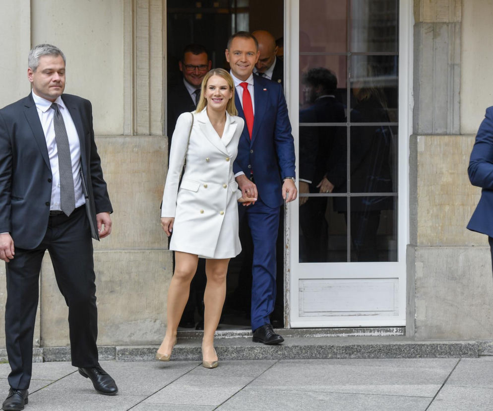 Marta Nawrocka w białej sukience o fasonie marynarki z złotymi guzikami i beżowych szpilkach, uśmiechnięta, wychodzi z budynku, trzymając za rękę mężczyznę w granatowym garniturze i czerwonym krawacie. O ocenach jej stylu przeczytasz na naszym portalu.