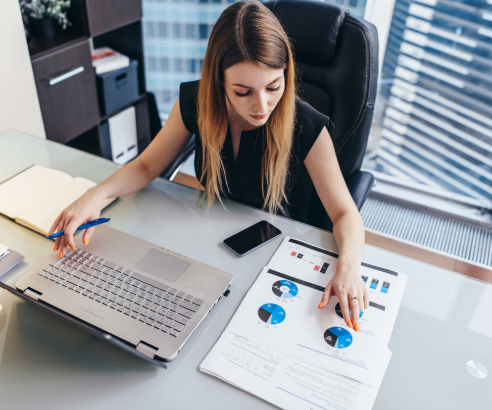 Kobieta pracująca przy biurku z laptopem i dokumentami z wykresami, odzwierciedlająca tematykę nowych przepisów pracy od 2026 roku. Analizuje dane finansowe i zmiany dotyczące etatów, staży oraz płac, co jest kluczowe dla firm, o czym przeczytasz na Super Biznes.