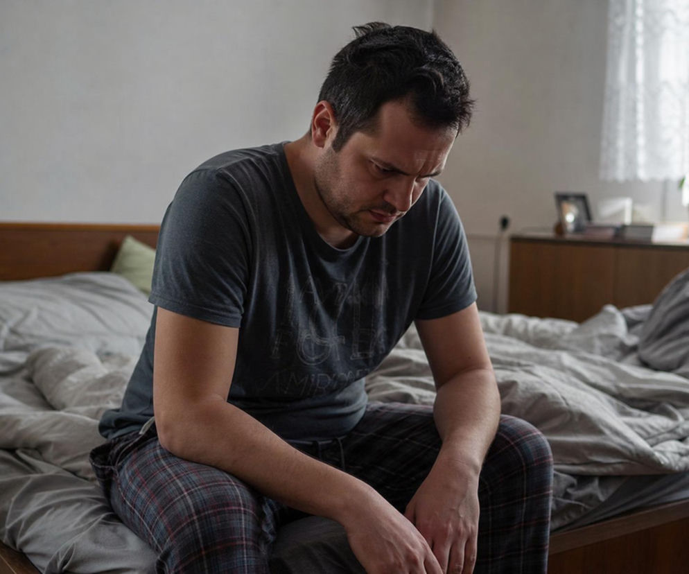 Mężczyzna zamyślony, siedzący na brzegu łóżka, w piżamie i szarej koszulce. Jego mina wyraża smutek i przygnębienie, co odzwierciedla emocje z historii o zdradzie, którą możesz przeczytać na naszym portalu. Obrazek oddaje poczucie winy i bezradności głównego bohatera.