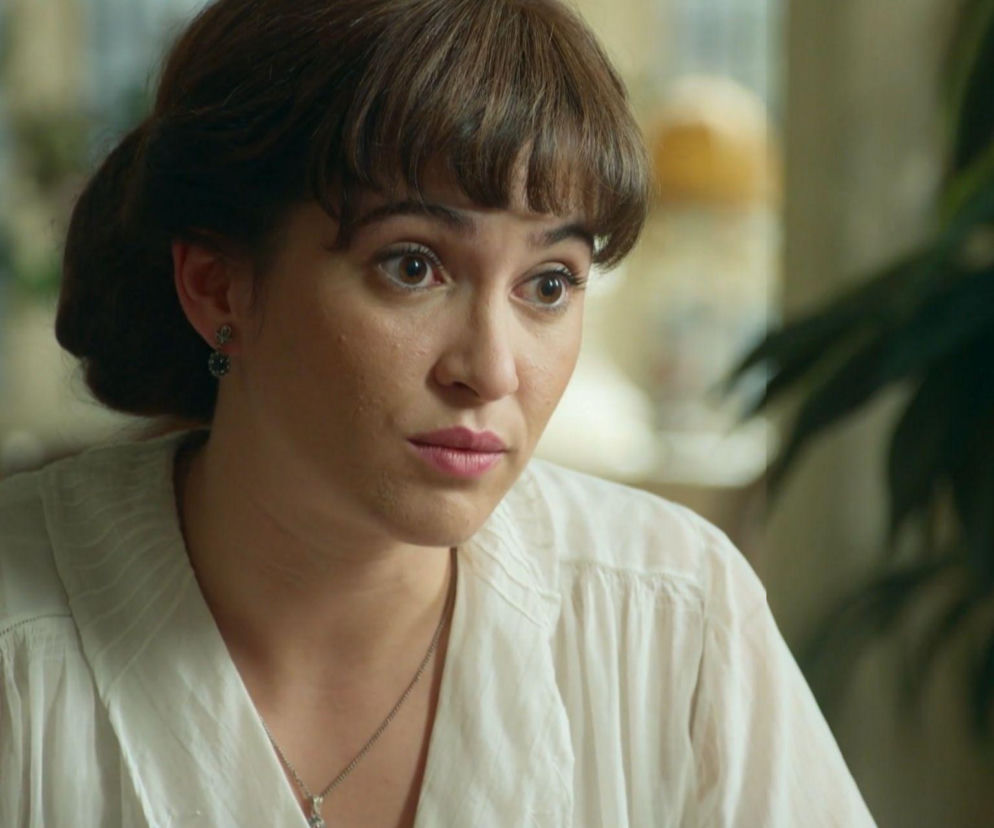 Bliskie ujęcie kobiety, Cataliny Luján z serialu La Promesa, z brązowymi włosami i grzywką, patrzącej w prawo z zaskoczeniem. Ma delikatny makijaż, jasne usta i kolczyki. Ubrana jest w białą, luźną bluzkę z kołnierzykiem i nosi naszyjnik. Więcej o losach bohaterki na Super Seriale.