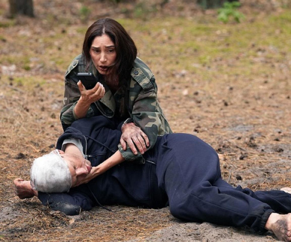 Celina (Orina Krajewska) w kurtce moro z brudnymi dłońmi klęczy przy wycieńczonym Łukaszu (Michał Rolnicki), który leży na ziemi w ciemnym dresie. Celina trzyma telefon przy uchu, wzywając pomoc. Zdjęcie ilustruje scenę z serialu „Barwy szczęścia”, o której można przeczytać na Super Seriale.