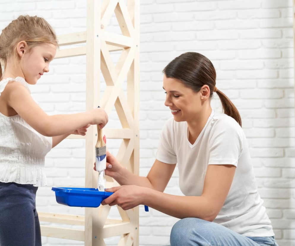 Uśmiechnięta mama z córką malują białą farbą drewniany regał. O wspieraniu dziecka w remoncie przeczytasz na SE.