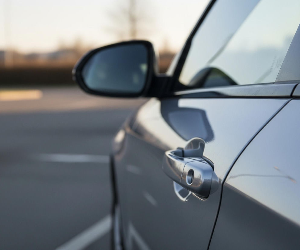 Na pierwszym planie, z prawej strony, znajduje się boczna część samochodu w kolorze srebrnym lub jasnoszarym, z widoczną klamką i fragmentem lusterka bocznego. Linie karoserii i okien są wyraźnie zarysowane, a powierzchnia odbija światło, tworząc refleksy. Lewa strona obrazu jest rozmyta i przedstawia ciemny asfalt z białymi liniami, sugerującymi miejsce parkingowe, oraz niewyraźne drzewa i budynki na horyzoncie pod jasnym, zachodzącym słońcem.