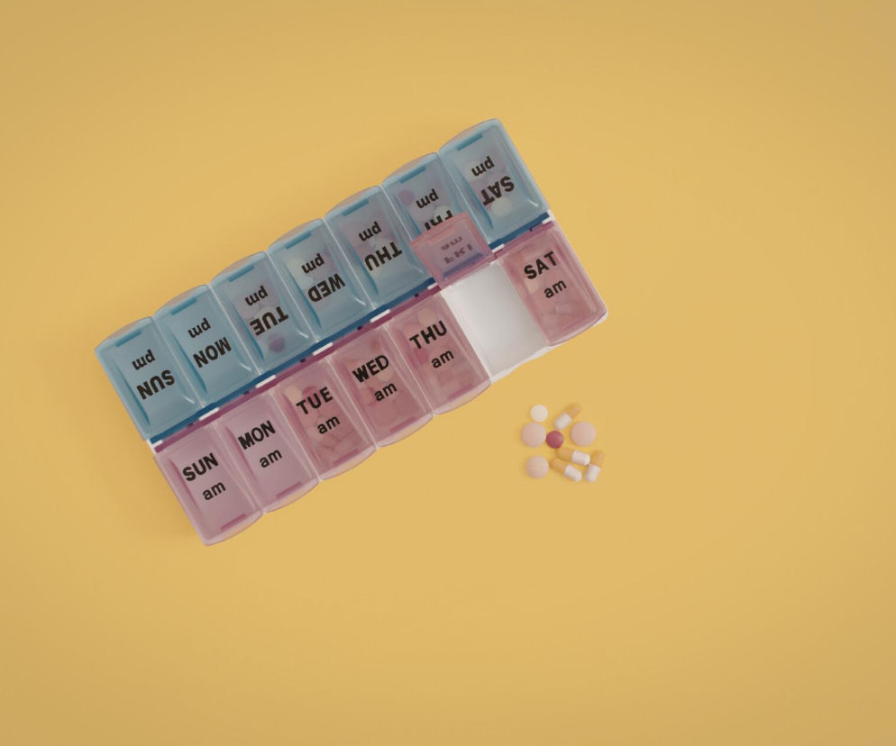 Organizer na tabletki w kolorach niebieskim i różowym, ułożony na żółtym tle. Obok leżą rozrzucone pigułki. Ilustracja symbolizuje regularne przyjmowanie suplementów, np. magnezu. Dowiedz się więcej o porze przyjmowania magnezu na Poradnik Zdrowie.