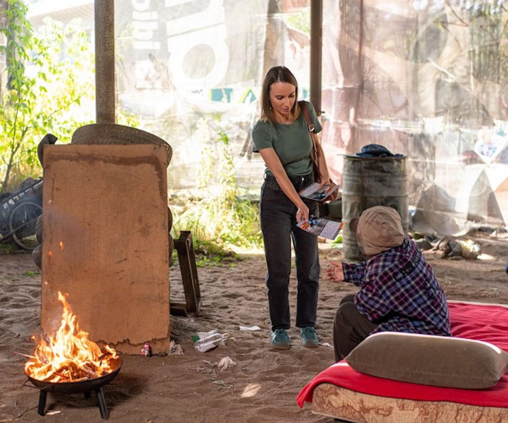 Ewa Jakubowicz jako Melka z serialu Pierwsza miłość rozmawia z bezdomnym, wręczając mu ulotki, w tle prowizoryczne schronienie i palenisko. Aktorka wciela się w postać, która angażuje się w pomoc osobom w kryzysie bezdomności, o czym przeczytasz na stronie Super Seriale.