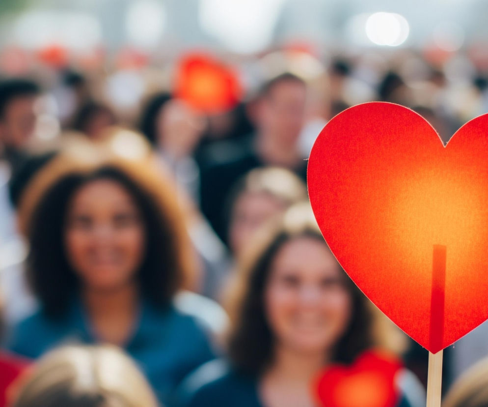 Czerwone serce na patyku, widoczne w prawym dolnym rogu, dominuje pierwszy plan obrazu. Jest ono wyraźnie oświetlone od tyłu, co tworzy jaśniejszy, pomarańczowy odcień w jego centrum. Za sercem rozciąga się rozmyty tłum ludzi, z których kilku trzyma podobne, mniejsze, czerwone serca. Osoby w pierwszym rzędzie, po lewej i prawej stronie serca, uśmiechają się, ich twarze są jednak rozmyte.