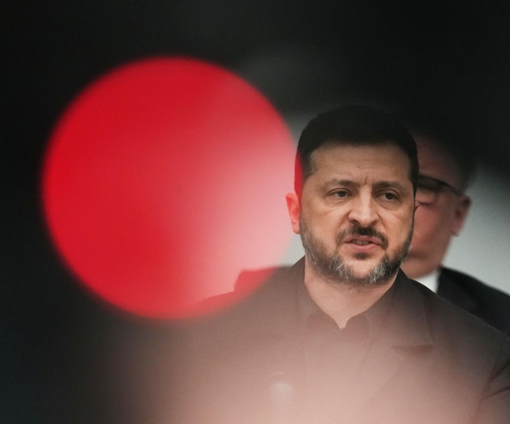 Prezydent Ukrainy, Wołodymyr Zełenski, ubrany w ciemną koszulę, podczas konferencji, z rozmytym, czerwonym światłem w tle. Widać, jak mówi do mikrofonu, co można przeczytać na naszym portalu.