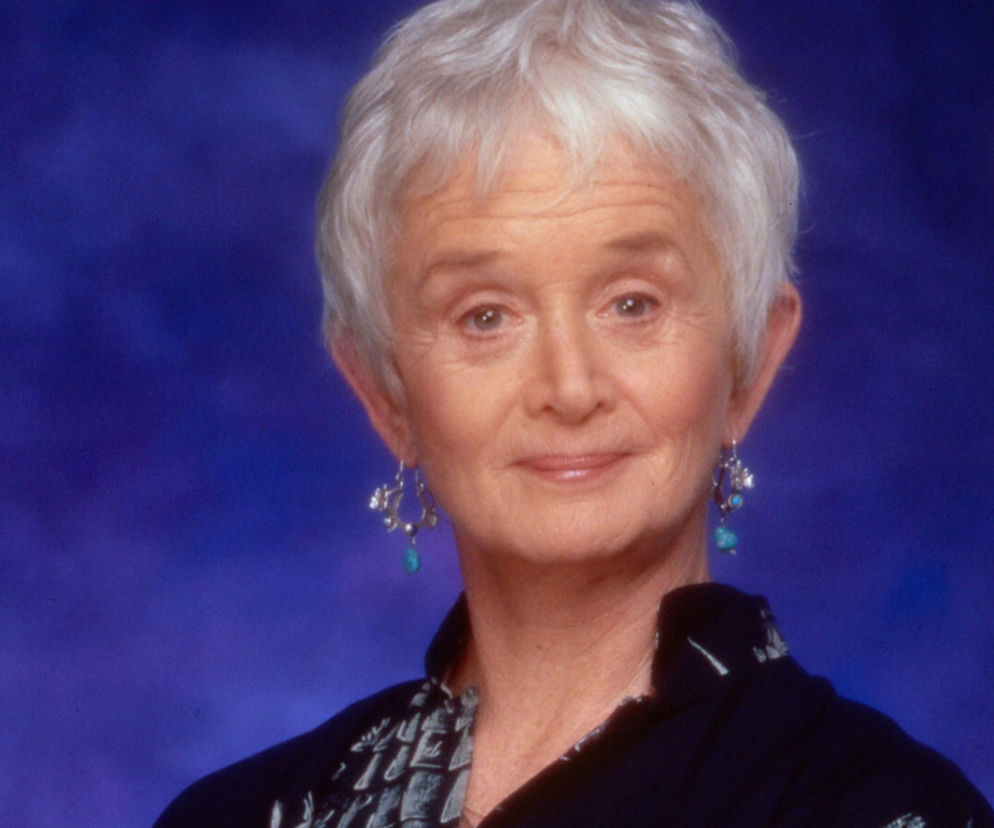 Aktorka Barbara Barrie z lekkim uśmiechem na tle niebieskiego tła. O jej śmierci przeczytasz na SE.