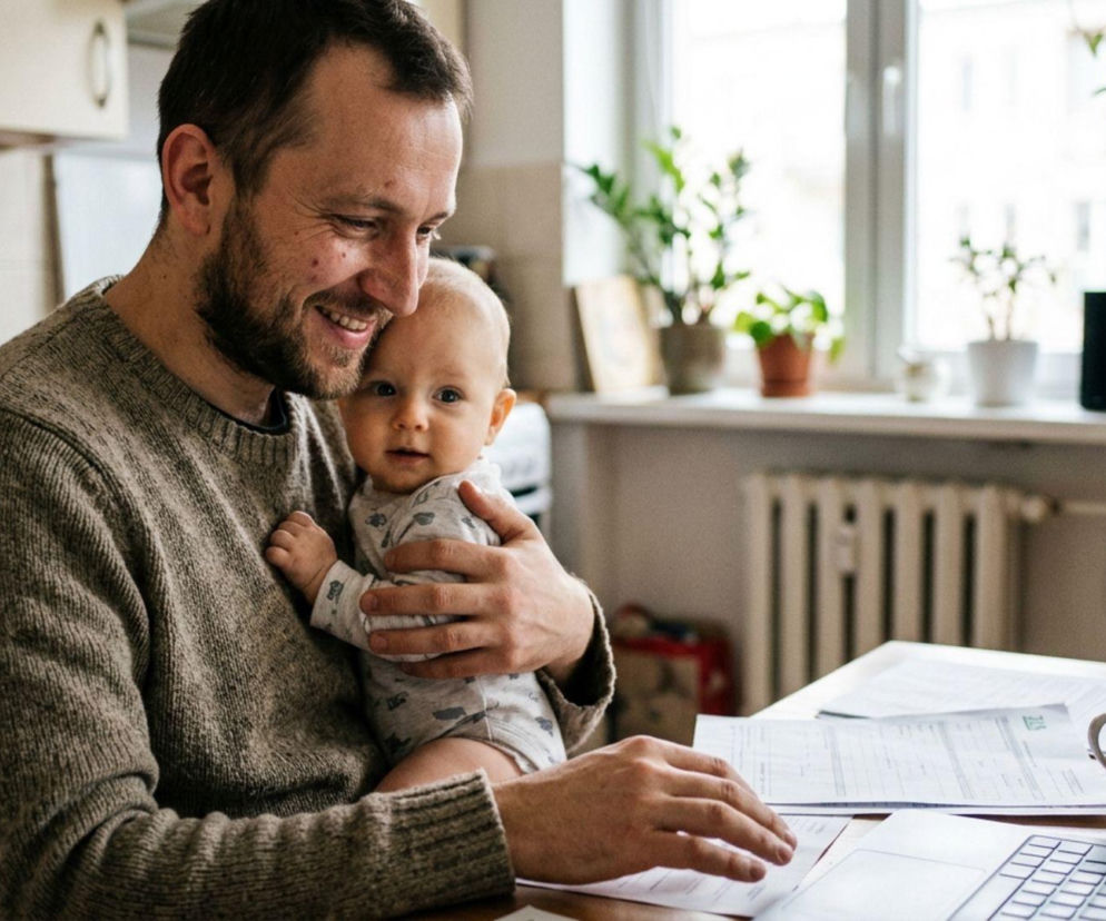 Szczęśliwy ojciec z uśmiechniętym niemowlęciem na rękach, siedzący przy stole z laptopem i dokumentami, co symbolizuje możliwość dłuższego urlopu dla ojca i budowanie więzi rodzinnych. Więcej o nowym trendzie przeczytasz na Super Biznes.