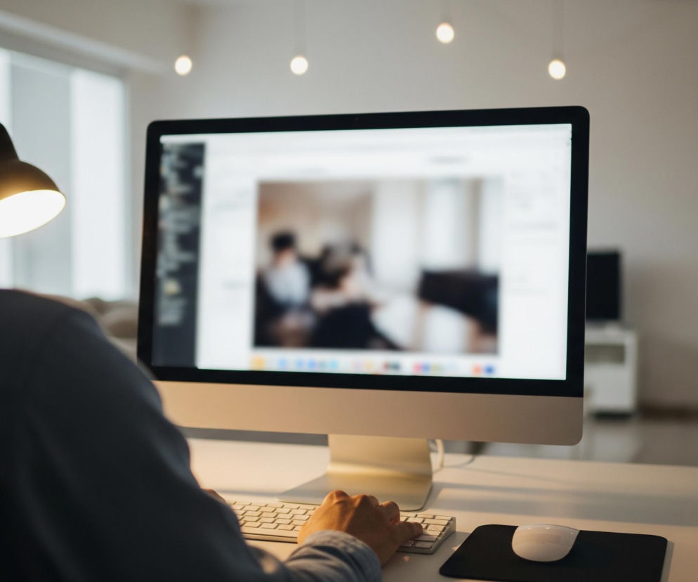 Mężczyzna w niebieskiej koszuli siedzi tyłem do widza, patrząc na monitor komputera. Jego prawa ręka spoczywa na białej klawiaturze, a obok, na czarnej podkładce, znajduje się biała mysz komputerowa. Na ekranie monitora widoczna jest rozmyta grafika przedstawiająca, prawdopodobnie, spotkanie lub rozmowę kilku osób. Na lewo od monitora stoi włączona lampka biurkowa, rzucająca ciepłe światło. W tle, w rozmyciu, widać białe ściany, okno, oraz rozproszone źródła światła na suficie.