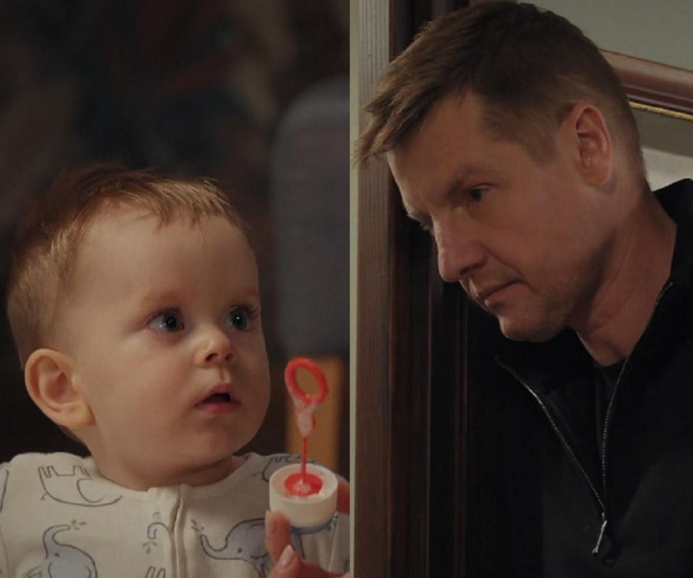 Mały Antoś z serialu M jak miłość z bujnymi rudymi włosami, ubrany w białe śpioszki w słonie, patrzy w górę, trzymając w ręku zabawkę do baniek mydlanych. Obok niego, Paweł (Rafał Mroczek) w czarnej bluzie, spogląda na dziecko. Więcej o ich losach na Super Seriale.