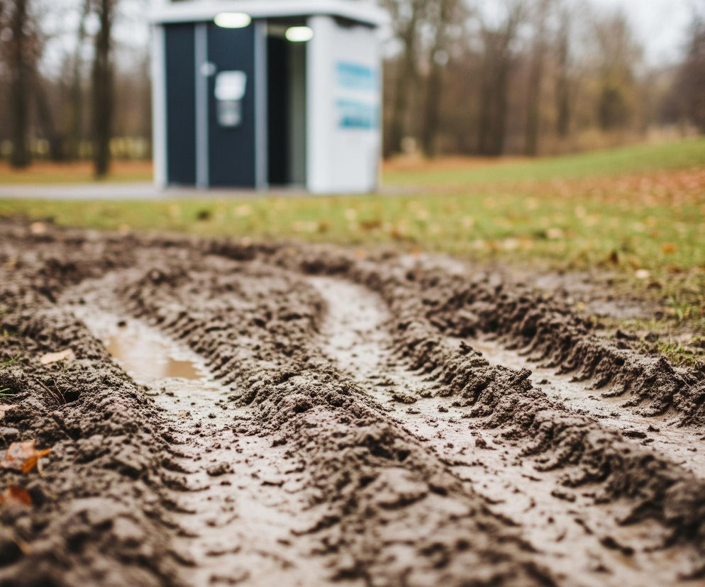 Na pierwszym planie widoczne są ślady opon pojazdu, głęboko wciśnięte w błotnistą, ciemnobrązową ziemię, z drobnymi kałużami wody. Dalej, po bokach i między koleinami, rośnie zielona trawa, gdzieniegdzie pokryta jesiennymi, brązowo-pomarańczowymi liśćmi. W tle znajduje się biało-czarny, prostokątny budynek z dwoma jasnymi prostokątnymi lampami na górze, stojący na zielonej trawie obok szarej ścieżki, a za nim rozciąga się park z licznymi drzewami o nagich, ciemnych gałęziach.