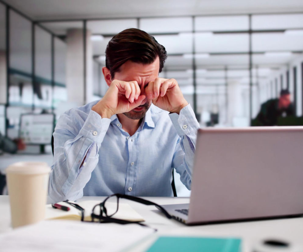 Mężczyzna w jasnej koszuli siedzący przy biurku w biurze, pociera zmęczone i piekące oczy. Laptop jest otwarty przed nim, a na blacie widoczne są też okulary i kubek. Ten obrazek ilustruje problem suchych oczu w klimatyzowanych pomieszczeniach, o czym możesz przeczytać na Poradnik Zdrowie.
