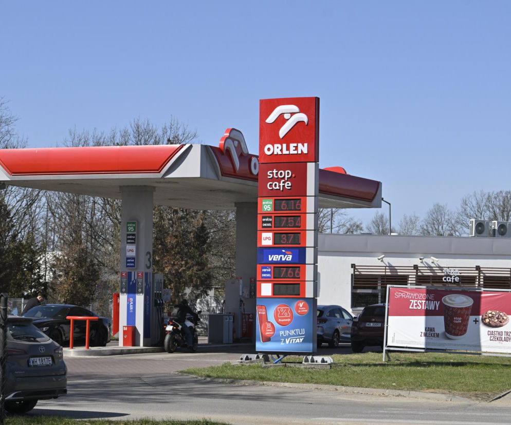 Stacja paliw Orlen z widocznym słupem cenowym, prezentującym aktualne ceny paliw, w tym Pb95, Pb98, LPG i olej napędowy. Samochody tankują na tle budynku Stop Cafe. Więcej o podwyżkach cen paliw na Super Biznes.