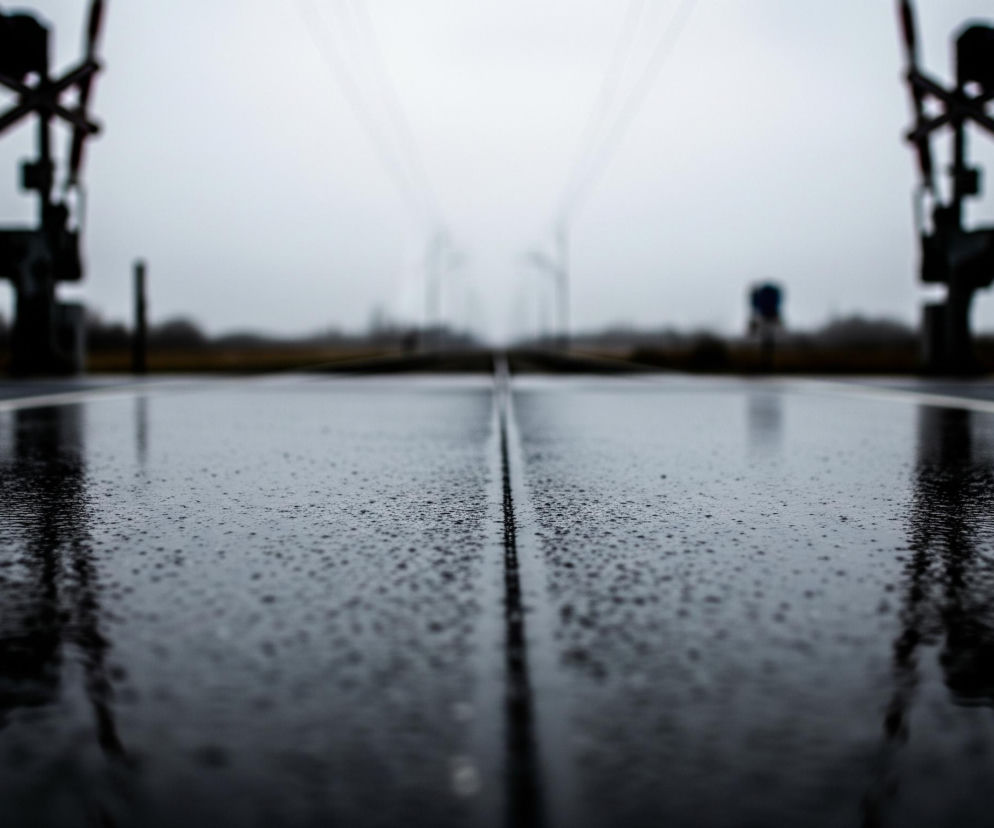 Głównym elementem obrazu jest mokra, ciemna nawierzchnia, prawdopodobnie asfaltowa, pokryta licznymi kropelkami wody, co sugeruje deszcz. Nawierzchnia rozciąga się od dołu obrazu, gdzie jest ostra i szczegółowa, w kierunku oddalonego, rozmytego horyzontu. Po obu stronach nawierzchni widać sylwetki rozmytych szlabanów kolejowych oraz sygnalizatorów, z jednym czerwonym światłem widocznym po prawej stronie. Tło stanowi zamglone, jasnoszare niebo, a na horyzoncie można dostrzec niewyraźne, ciemne obiekty i linię drzew.