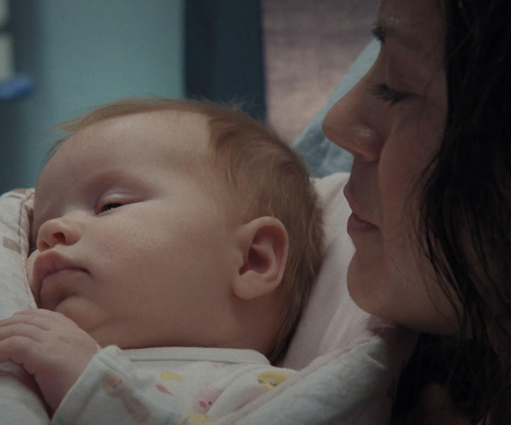 Noworodek Zosia o rudych włoskach, śpiący w szpitalnym łóżeczku obok swojej matki, Kasi (Paulina Lasota) z serialu M jak miłość. Matka z długimi, ciemnymi włosami, patrzy z czułością na dziecko. Artykuł o tym, dlaczego Zosia nigdy nie pozna prawdziwego ojca znajdziesz na Super Seriale.