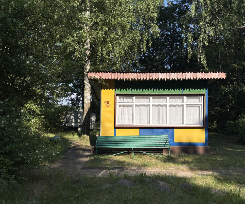 Drewniany domek letniskowy nr 18 w ośrodku nad Pilicą, utrzymany w kolorach żółtym, niebieskim i zielonym, z zieloną ławką przed wejściem. To architektura lat 70. XX wieku. Więcej o domkach można przeczytać na Architektura Murator Plus.