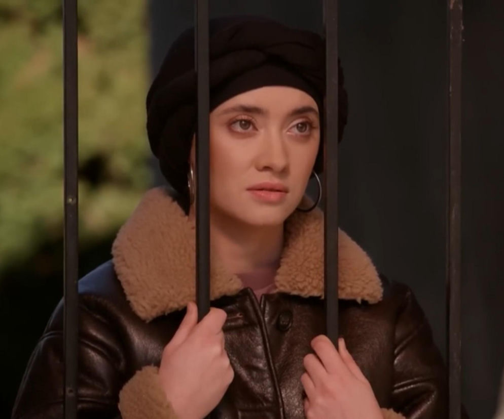 Patrycja Franczak jako Angelika w czarnym turbanie i brązowej kurtce z futerkiem, zmartwiona trzymająca kraty. Emocjonalny kadr z Pierwszej miłości, zapowiadający dramatyczne wydarzenia po wakacjach, o których przeczytasz na Super Seriale.