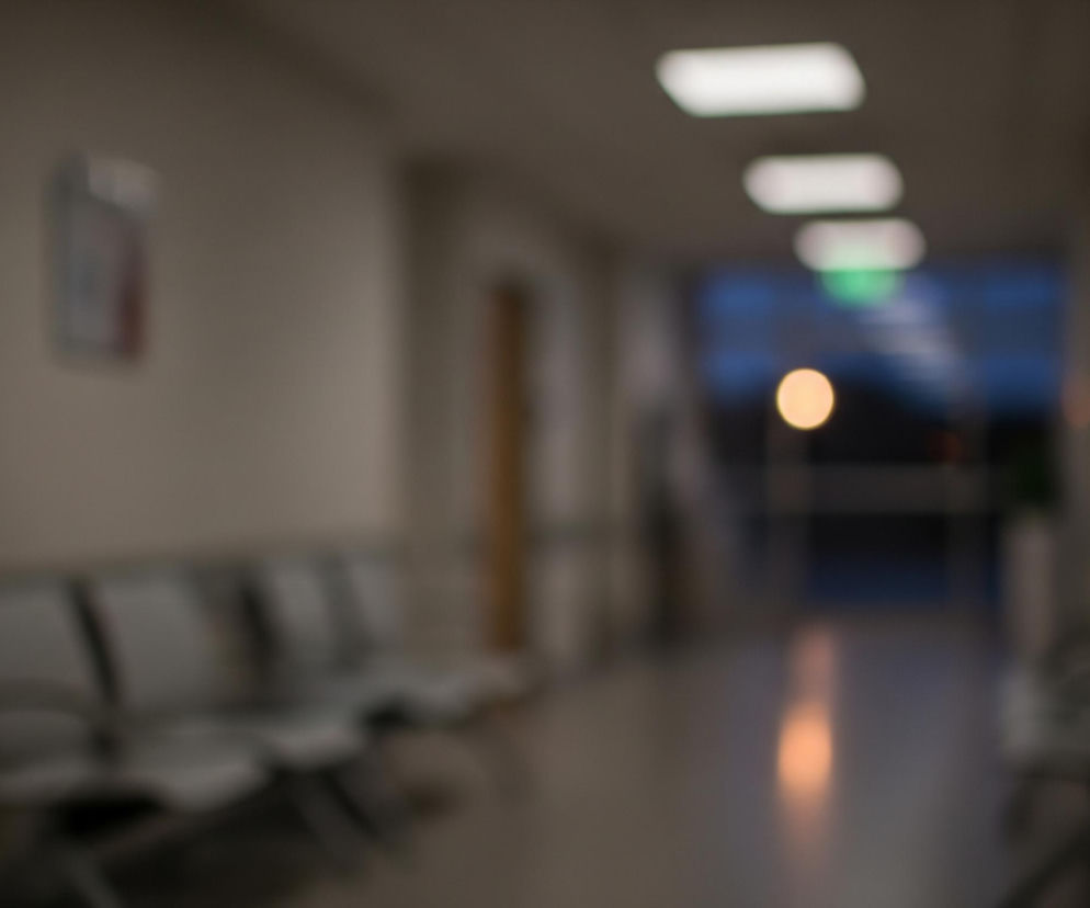 Długi korytarz z kremowymi ścianami rozciąga się w głąb obrazu, gdzie na końcu widać jaśniejsze, niebieskawe światło z zewnątrz oraz wyraźną, pomarańczową plamę świetlną. Po lewej stronie korytarza ustawione są rzędy krzeseł z metalowymi ramami i jasnymi siedziskami, a na ścianie wisi prostokątny, rozmazany obrazek i ciemny, kwadratowy element. Sufit jest szary z widocznymi, podłużnymi, białymi lampami, których intensywność maleje w oddali. Na podłodze odbija się pomarańczowe światło.