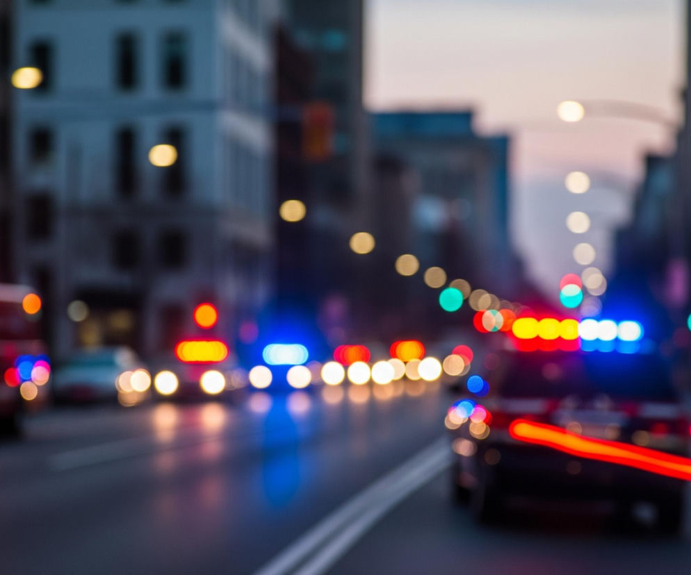 Uliczna scena nocą, rozmyta w efekcie bokeh, zdominowana przez jasne, kolorowe światła. Po lewej stronie widać rozmyty czerwony pojazd, prawdopodobnie ambulans lub wóz strażacki, z jaskrawoczerwonymi światłami ostrzegawczymi na dachu oraz żółtymi światłami z przodu. Po prawej stronie drogi znajduje się ciemniejszy pojazd, również z niebieskimi i czerwonymi światłami ostrzegawczymi na górze oraz dwoma długimi, poziomymi czerwonymi smugami światła z tyłu, sugerującymi ruch. Tło wypełniają rozmyte budynki i liczne, okrągłe światła uliczne oraz sygnalizacyjne w odcieniach żółci, zieleni, czerwieni i błękitu, tworzącą dynamiczną i enigmatyczną atmosferę miejskiego krajobrazu.
