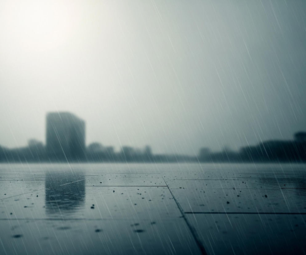 Ulewny deszcz pada na mokrą powierzchnię bruku w mieście. Głębia ostrości skupia się na pierwszym planie, gdzie widoczne są kafelki chodnika z licznymi kroplami wody, z których niektóre tworzą drobne kałuże. W tle rozmazane są ciemne sylwetki budynków pod szarym, zachmurzonym niebem, a widoczna jest też jaśniejsza plama, prawdopodobnie słońca przebijającego się przez chmury. Cały obraz utrzymany jest w zimnych odcieniach szarości i błękitu, co potęguje wrażenie ponurej i deszczowej pogody.