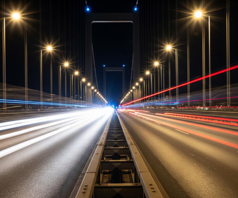 Droga na moście oświetlona jest licznymi lampami ulicznymi z obu stron, z długimi, promienistymi efektami świetlnymi, co tworzy intensywny blask na nocnym niebie. Po lewej stronie jezdni widoczne są białe smugi światła, oznaczające ruch pojazdów w kierunku patrzącego, natomiast po prawej stronie dominują czerwone smugi świateł, symbolizujące pojazdy oddalające się od obserwatora. W centrum kompozycji, w oddali, widać jasne światła świateł drogowych i pojedyncze niebieskie punkty, prawdopodobnie dekoracje mostu. Konstrukcja mostu, z jego pylonami i linami, jest zarysowana ciemno na tle czarnego nieba.